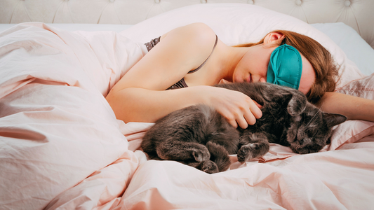 Person sleeping in bed while hugging a gray cat, wearing a teal sleep mask.